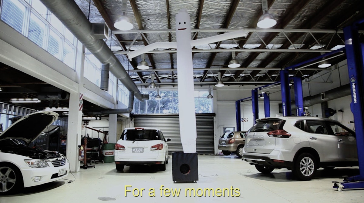 Crystal, Elisabeth's inflatable waving man is standing upright in a luxury car dealership workshop. There are yellow subtitles on the image reading "for a few moments"